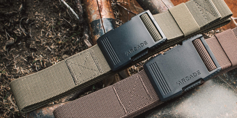 Two Arcade Belts, one khaki and one brown, crafted with stretch webbing and black plastic buckles labeled ARCADE, are placed side by side on a rustic metal surface outdoors.