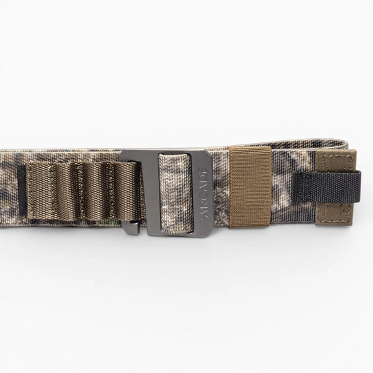 The Hardware Alu by Arcade Belts is a camouflage-patterned travel belt with a matte black plastic buckle, stretch webbing, elastic loops, and brown and black accents, shown coiled on a white background.