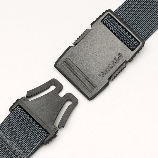 A close-up of a black plastic buckle on a dark woven strap, partially unclipped, with ARCADE engraved. The Arcade Belts Hardware belt stands out for its superior strength against a plain white background.