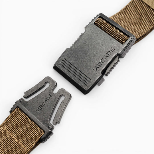 A close-up of the Arcade Belts Hardware, a brown elastic stretch belt with a black plastic buckle—partially unclasped and set against a white background. The ARCADE logo is visible on both parts of the low-profile buckle.