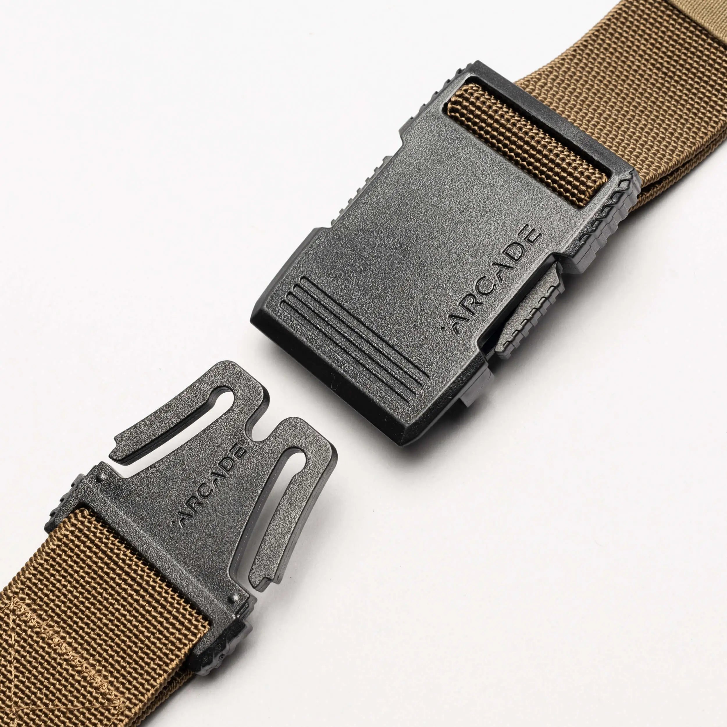 A close-up of the Arcade Belts Hardware, a brown elastic stretch belt with a black plastic buckle—partially unclasped and set against a white background. The ARCADE logo is visible on both parts of the low-profile buckle.