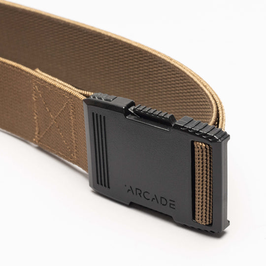 A close-up of the Arcade Belts Hardware, a tan elastic stretch belt with a low-profile black plastic buckle engraved with ARCADE, laid out on a plain white background.
