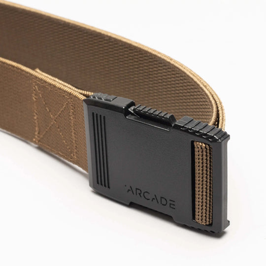 A close-up of the Arcade Belts Hardware, a tan elastic stretch belt with a low-profile black plastic buckle engraved with ARCADE, laid out on a plain white background.