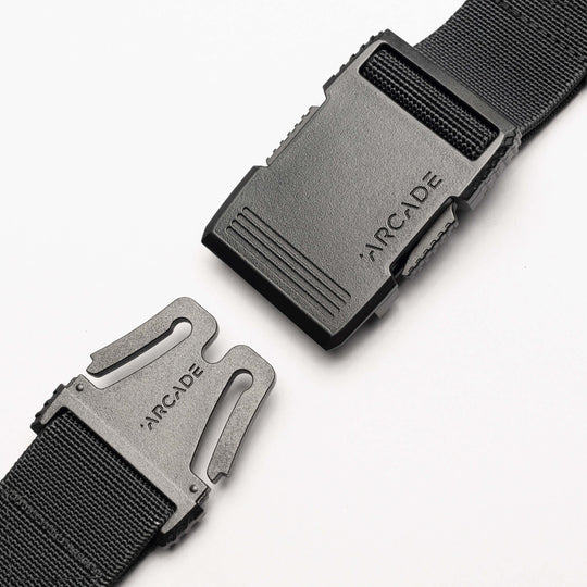 Close-up of the Arcade Belts Hardware black stretch belt with a plastic buckle, partially unclasped. Both buckle pieces are engraved with ARCADE. The sleek, low profile design is highlighted against a plain white background.