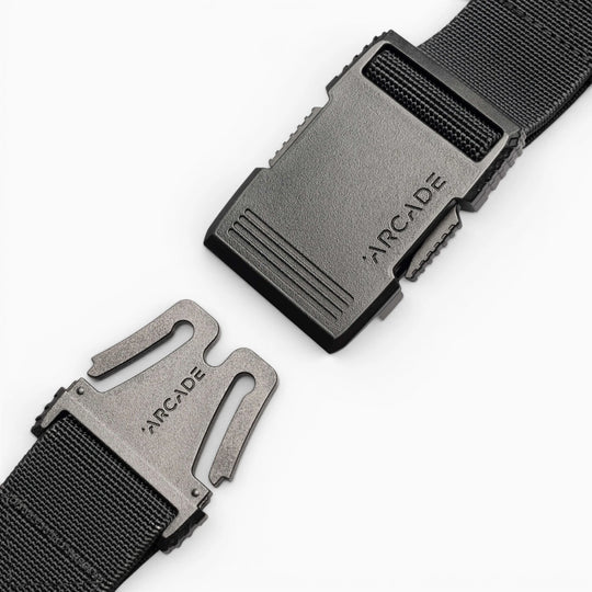 Close-up of the Arcade Belts Hardware black stretch belt with a plastic buckle, partially unclasped. Both buckle pieces are engraved with ARCADE. The sleek, low profile design is highlighted against a plain white background.