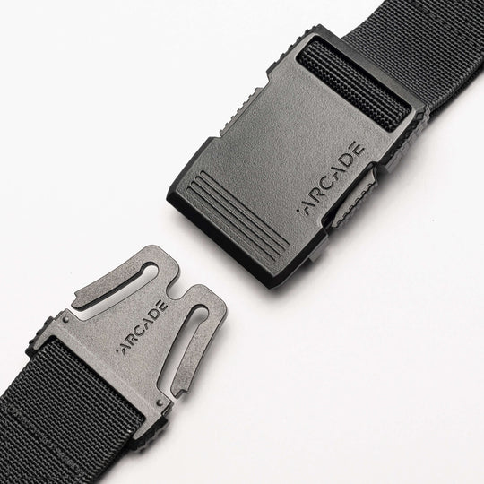 A close-up of the Arcade Belts Utility Long Kit, a black elastic stretch belt with a plastic buckle showing the brand name engraved on both clasp and buckle, displayed against a white background.