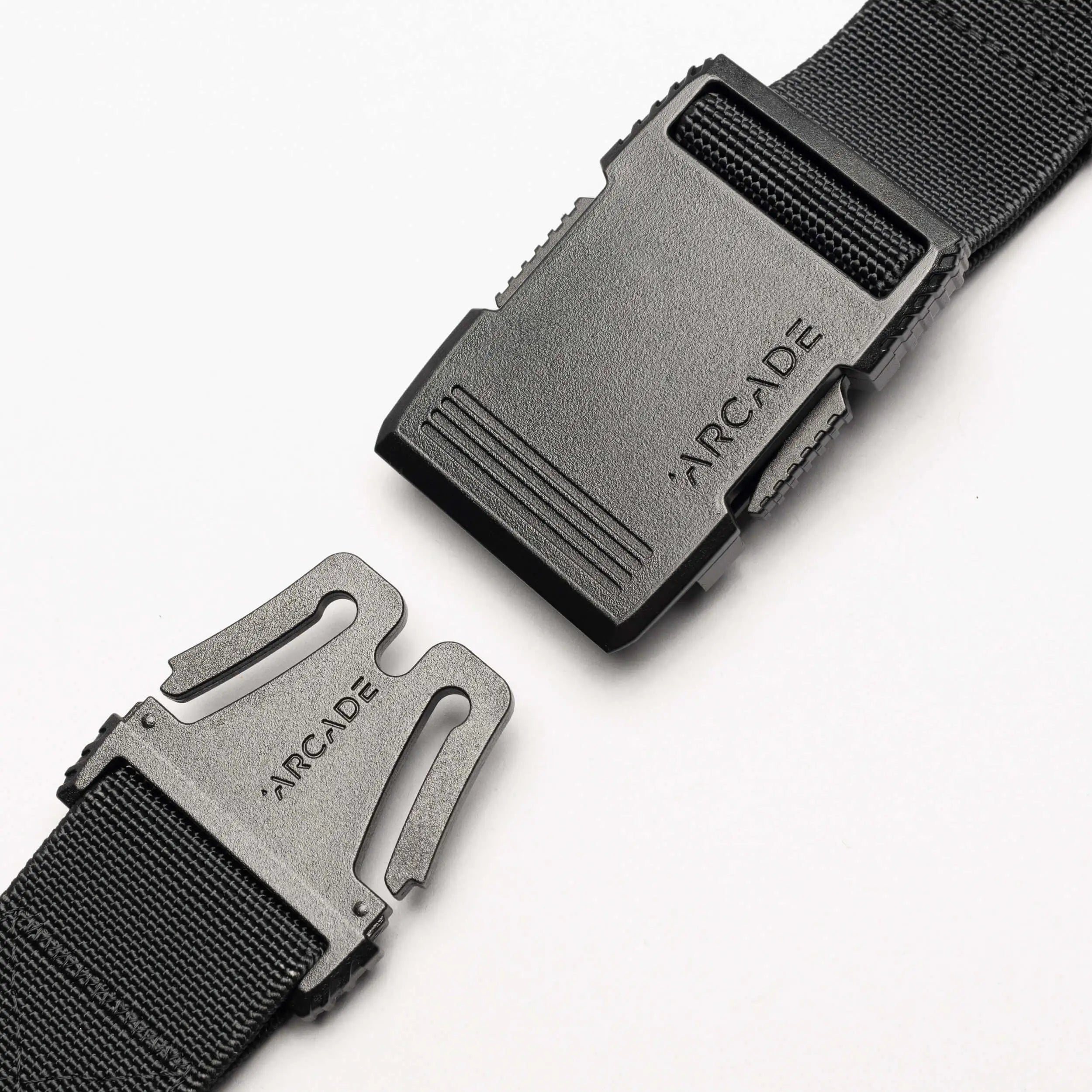 Close-up of the Arcade Belts Hardware black stretch belt with a plastic buckle, partially unclasped. Both buckle pieces are engraved with ARCADE. The sleek, low profile design is highlighted against a plain white background.