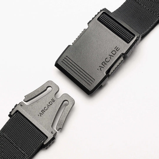 Close-up of the Arcade Belts Hardware black stretch belt with a plastic buckle, partially unclasped. Both buckle pieces are engraved with ARCADE. The sleek, low profile design is highlighted against a plain white background.