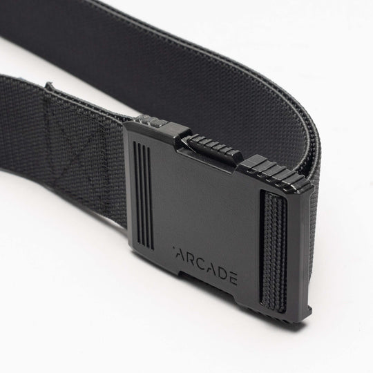 A close-up of the Arcade Belts Hardware: a black elastic stretch belt with a rectangular black plastic buckle engraved with “ARCADE,” shown against a white background.