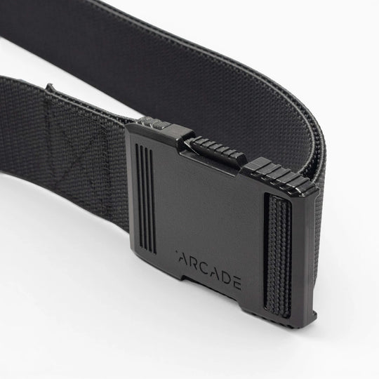 A close-up of the Arcade Belts Hardware: a black elastic stretch belt with a rectangular black plastic buckle engraved with “ARCADE,” shown against a white background.
