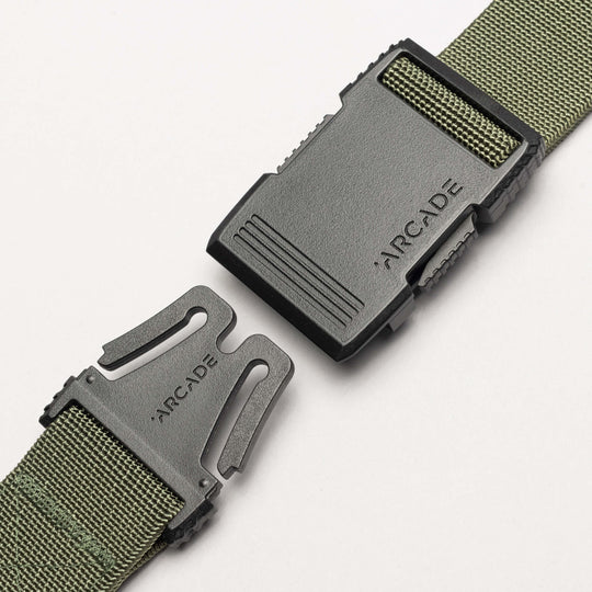 A close-up of the Arcade Belts Hardware, a green woven travel belt with stretch webbing and a black plastic buckle branded ARCADE on both halves, partially unfastened on a white background.
