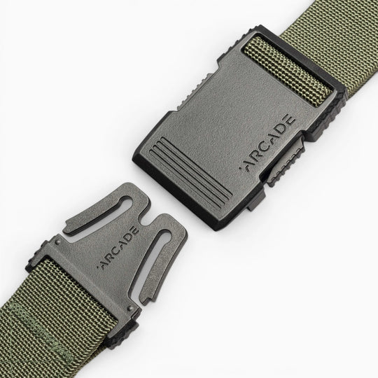 A close-up of the Arcade Belts Hardware, a green woven travel belt with stretch webbing and a black plastic buckle branded ARCADE on both halves, partially unfastened on a white background.