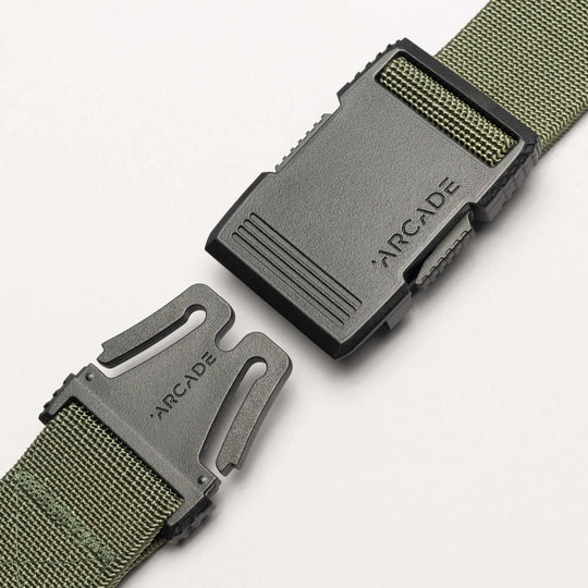 A close-up of the Arcade Belts Hardware, a green woven travel belt with stretch webbing and a black plastic buckle branded ARCADE on both halves, partially unfastened on a white background.