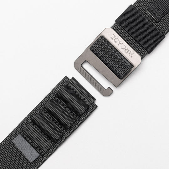 A close-up of Arcade Belts Hardware Alu, a black stretch webbing strap with a metal buckle and loop system, slightly detached on a white background. The low-profile belt features stitched rectangles and an ARCDE-engraved buckle.