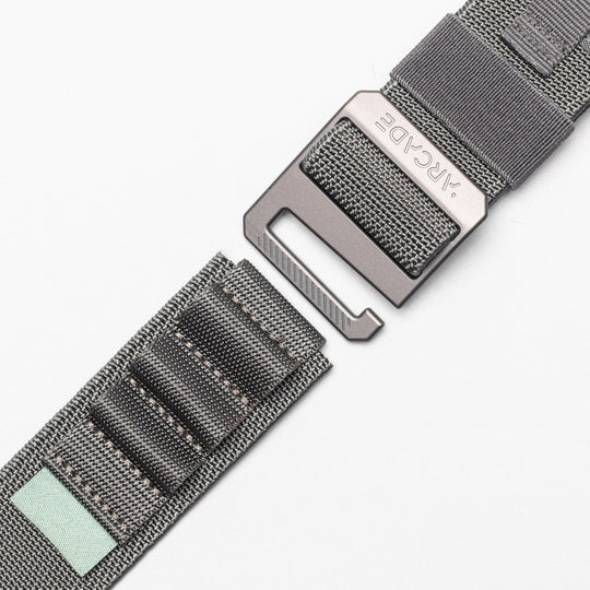 A close-up of the Arcade Belts Hardware Alu: a gray woven elastic stretch belt with stitched segments, a metal buckle, and a small light green fabric patch on one end, shown against a white background.