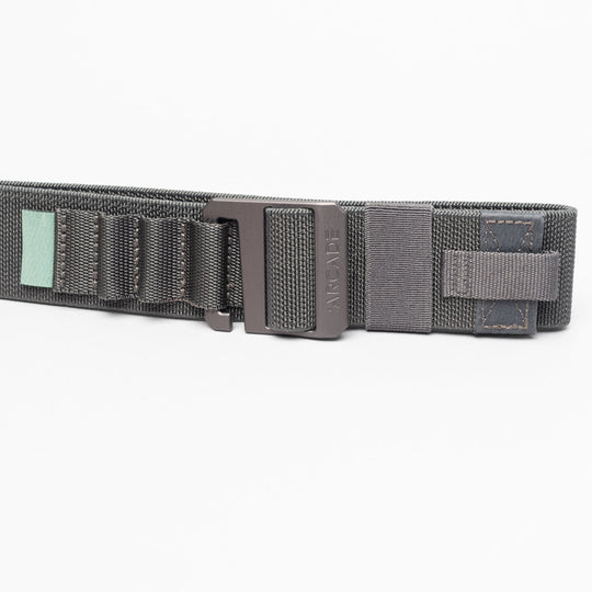 A close-up of the Arcade Belts Hardware Alu, a dark gray elastic stretch belt with a black metal buckle, stitched loops, and a small green tag, shown on a white background.