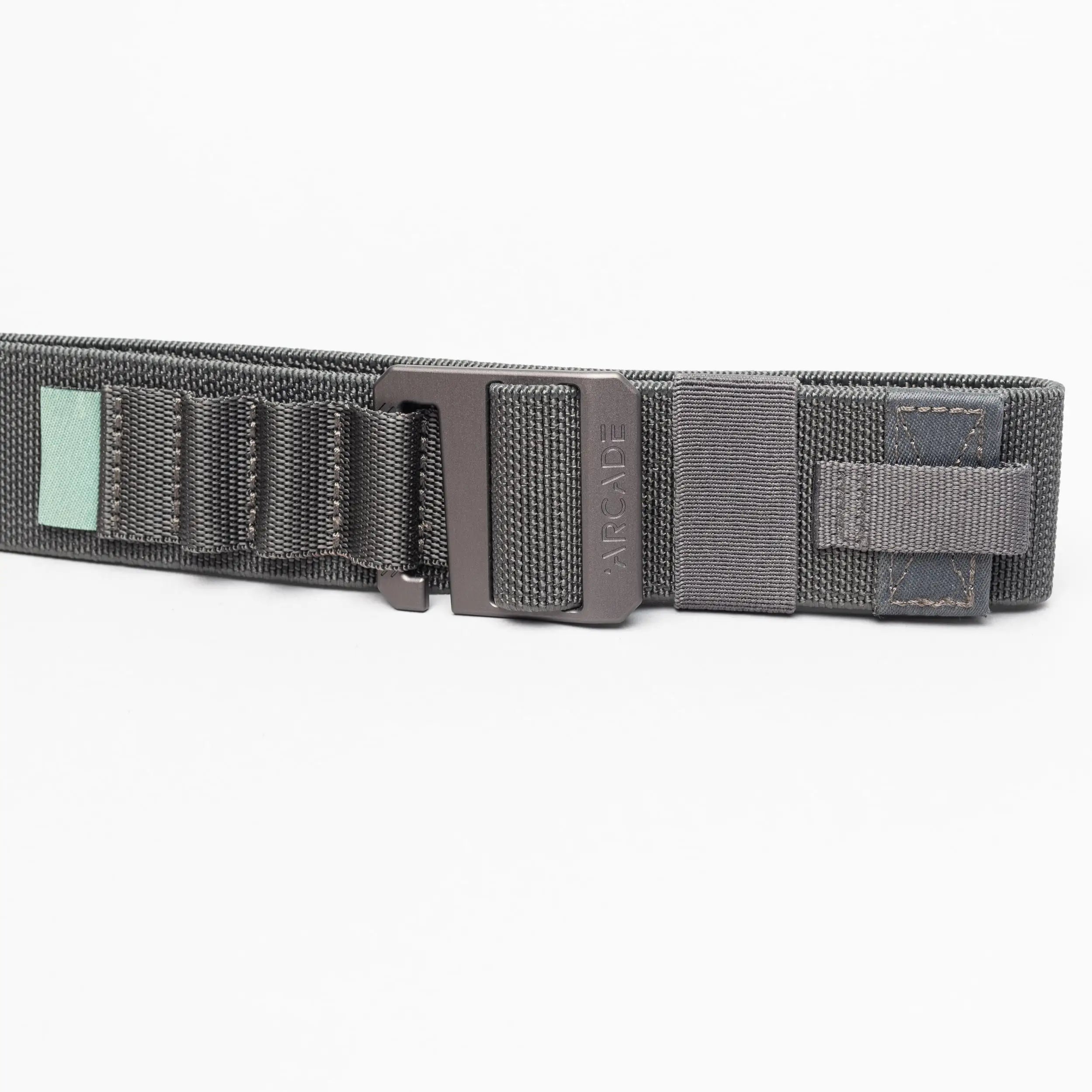 A close-up of the Arcade Belts Hardware Alu, a dark gray elastic stretch belt with a black metal buckle, stitched loops, and a small green tag, shown on a white background.