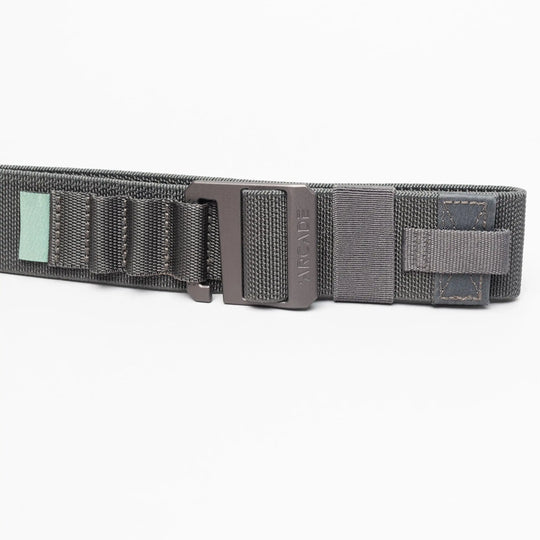 A close-up of the Arcade Belts Hardware Alu, a dark gray elastic stretch belt with a black metal buckle, stitched loops, and a small green tag, shown on a white background.
