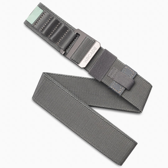 The Arcade Belts Hardware Alu is a grey woven stretch belt with a metal buckle, black accent stitching, and a mint green fabric tab at the end, displayed on a white background.