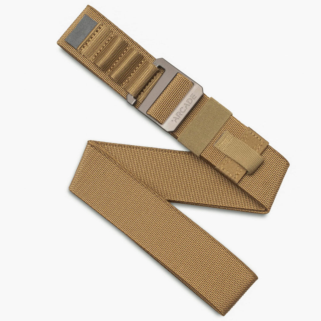 A tan-colored nylon belt with a rectangular metallic buckle. The belt is partially folded, showing its durable, textured fabric and stitched detailing. The buckle has engraved text and a modern, functional design.