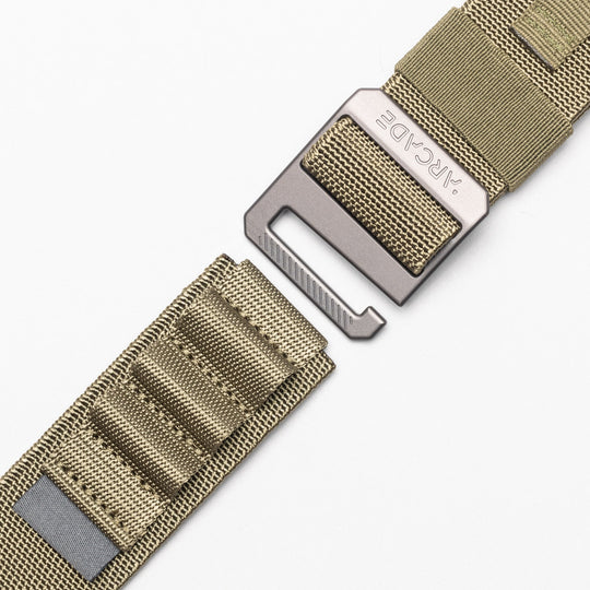 Close-up of a tan nylon Arcade Belts Hardware Alu belt with a rectangular metal buckle engraved “ARCADE.” The stretch webbing strap features stitched loops and a reflective strip, displayed on a white background.