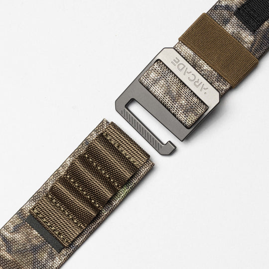 A close-up of the Hardware Alu belt by Arcade Belts, featuring a camouflage stretch webbing and a metal buckle, partially unfastened, displayed against a plain white background.