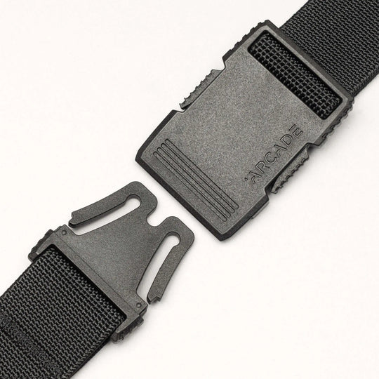 A close-up of a black plastic buckle on the Arcade Belts Hardware strap shows its two ends separated, with "ARCADE" embossed on the top piece. This belt offers superior strength against a clean white background.