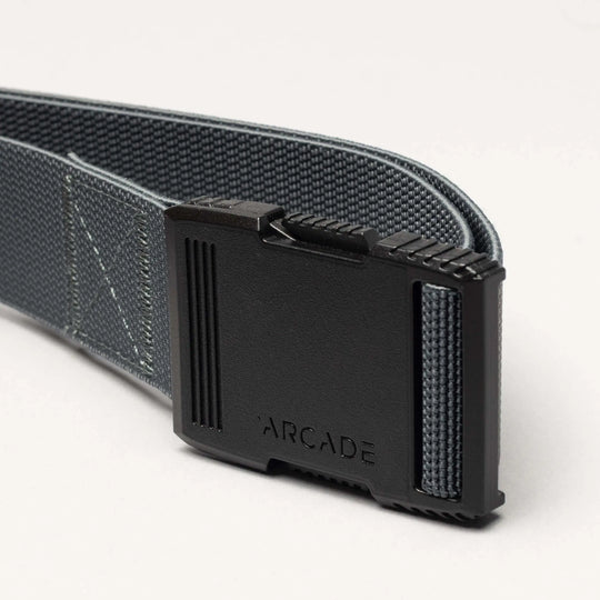 A close-up of the gray Arcade Belts Hardware belt highlights its superior strength and a rectangular black plastic buckle engraved with "ARCADE," set against a plain light-colored background.