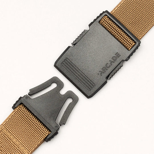 Close-up of a tan woven Arcade Belts Hardware belt featuring a black plastic buckle embossed with "ARCADE." The partially open buckle reveals its strong clasping mechanism.