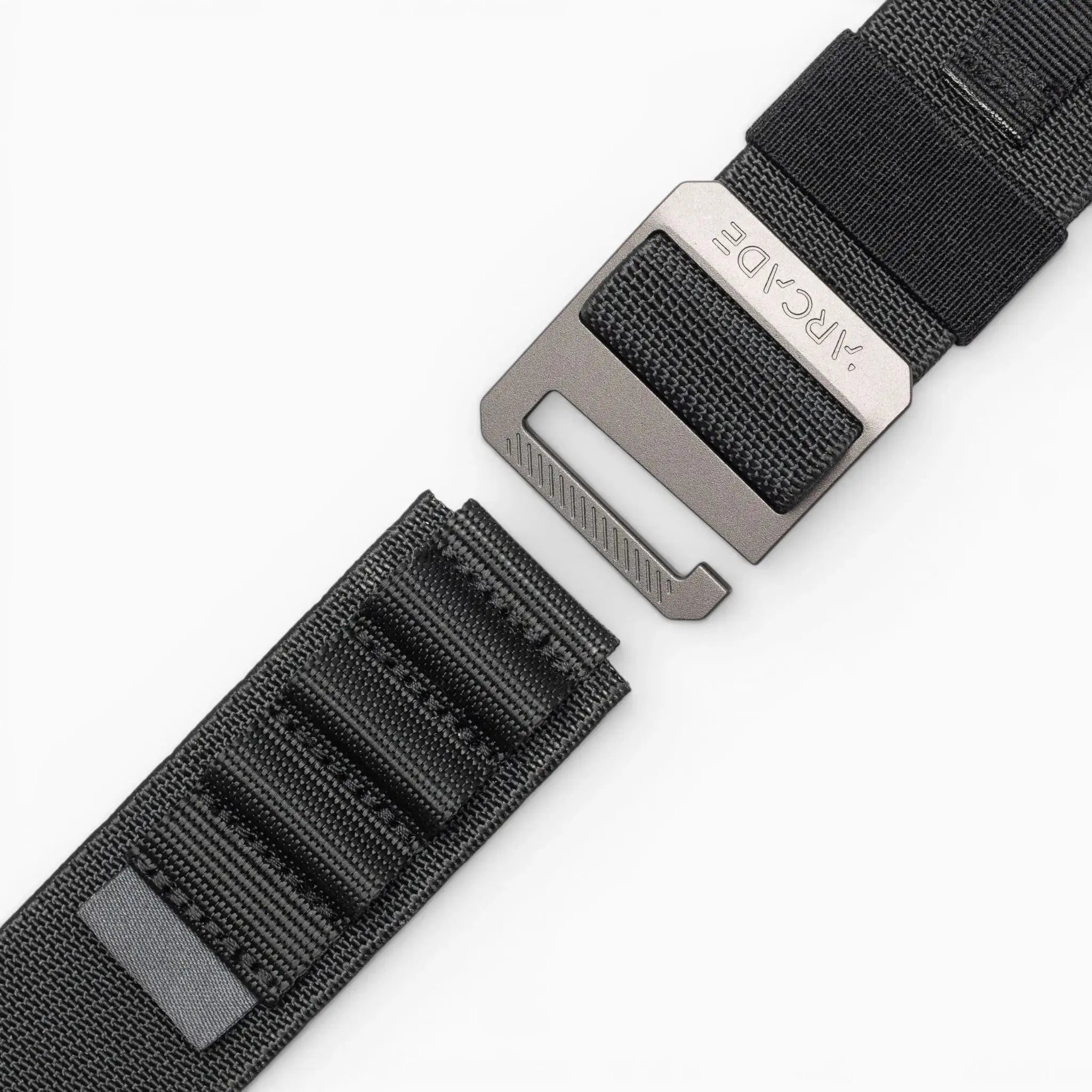 A close-up of Arcade Belts Hardware Alu, a black stretch webbing strap with a metal buckle and loop system, slightly detached on a white background. The low-profile belt features stitched rectangles and an ARCDE-engraved buckle.