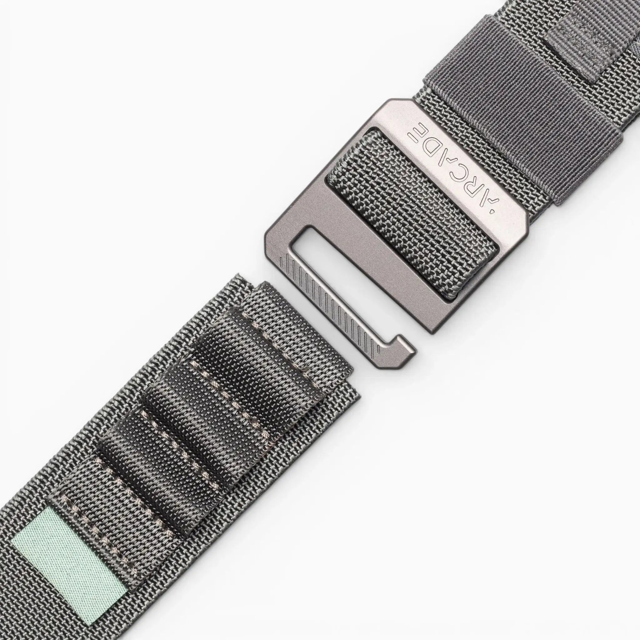 A close-up of the Arcade Belts Hardware Alu: a gray woven elastic stretch belt with stitched segments, a metal buckle, and a small light green fabric patch on one end, shown against a white background.