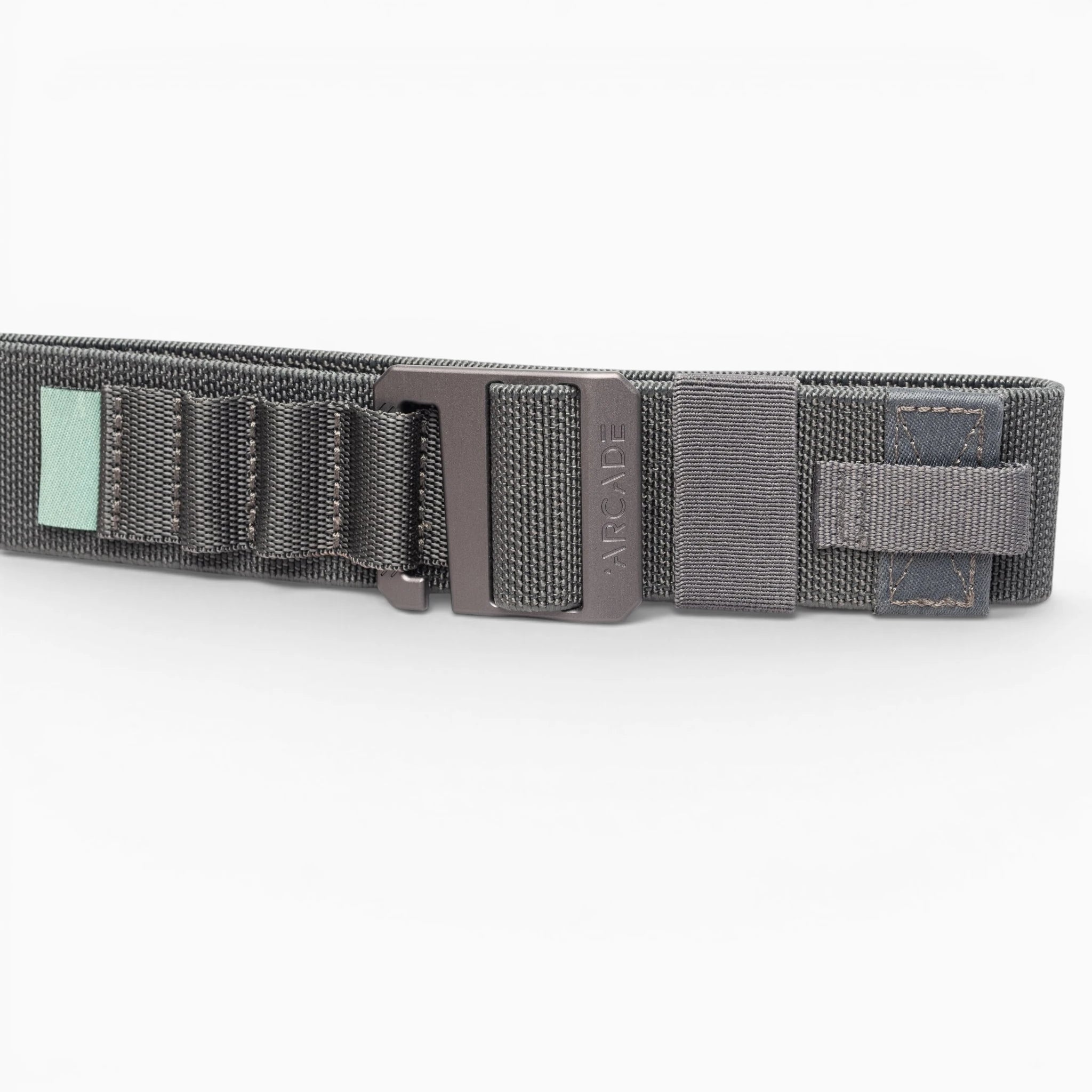 A close-up of the Arcade Belts Hardware Alu, a dark gray elastic stretch belt with a black metal buckle, stitched loops, and a small green tag, shown on a white background.