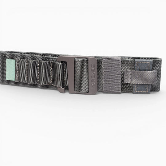 A close-up of the Arcade Belts Hardware Alu, a dark gray elastic stretch belt with a black metal buckle, stitched loops, and a small green tag, shown on a white background.