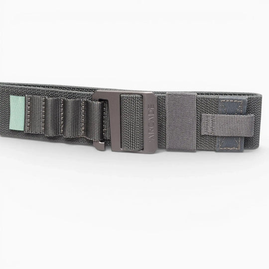 A close-up of the Arcade Belts Hardware Alu, a dark gray elastic stretch belt with a black metal buckle, stitched loops, and a small green tag, shown on a white background.