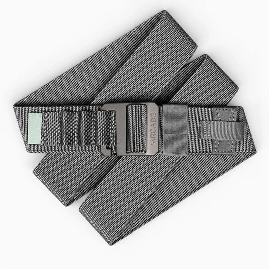 The Arcade Belts Hardware Alu is a gray fabric belt with stretch webbing, a black buckle engraved with Arcade, reinforced stitching, and a small rectangular patch near the buckle. Its neatly coiled and partially folded.