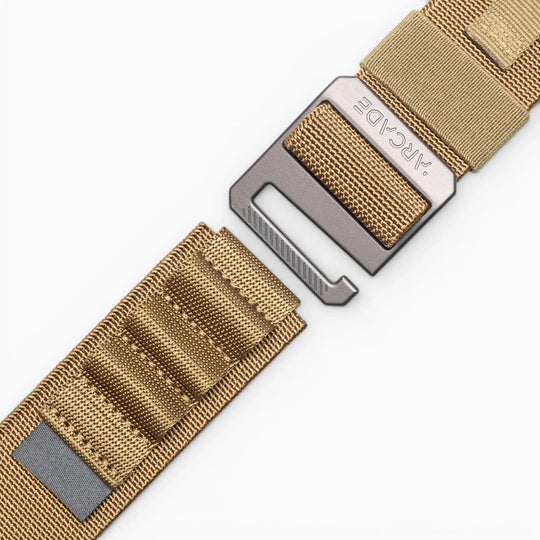 A close-up of the tan nylon Arcade Belts Hardware Alu belt with a metal buckle, set against a white background; the buckle and strap are partially separated.