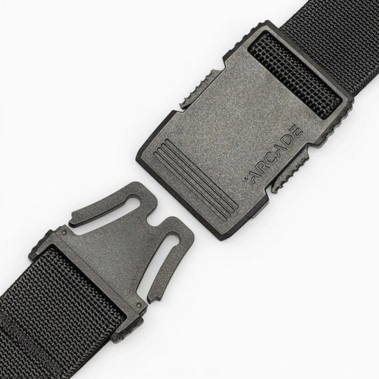 A close-up of a black plastic buckle on the Arcade Belts Hardware strap shows its two ends separated, with "ARCADE" embossed on the top piece. This belt offers superior strength against a clean white background.