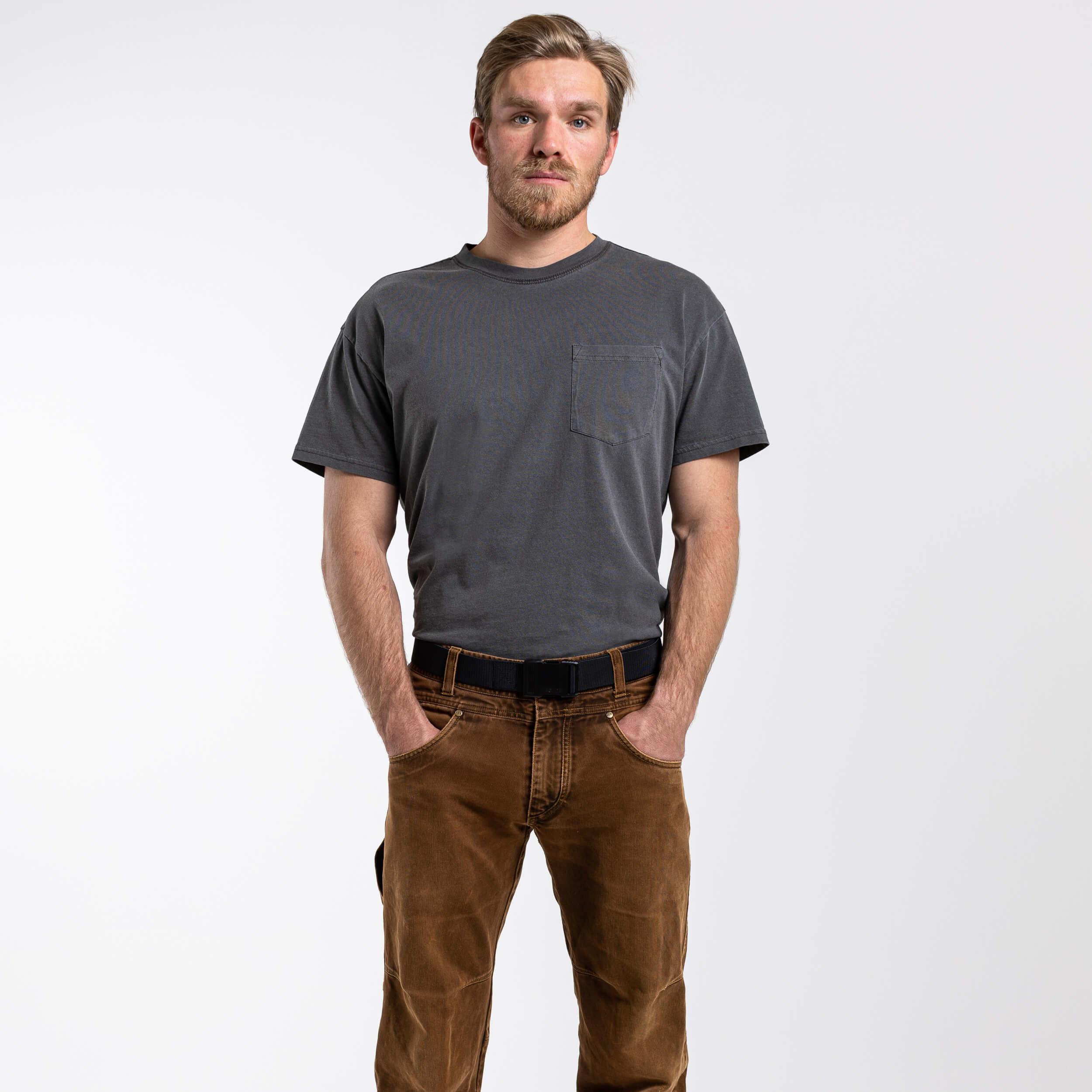 A light-haired, bearded man stands against a white background with his hands in his pockets, wearing a gray t-shirt, brown pants, and an Arcade Belts Midnight Long Kit belt as he looks at the camera.