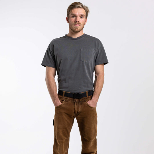 A light-haired, bearded man stands against a white background with his hands in his pockets, wearing a gray t-shirt, brown pants, and an Arcade Belts Midnight Long Kit belt as he looks at the camera.