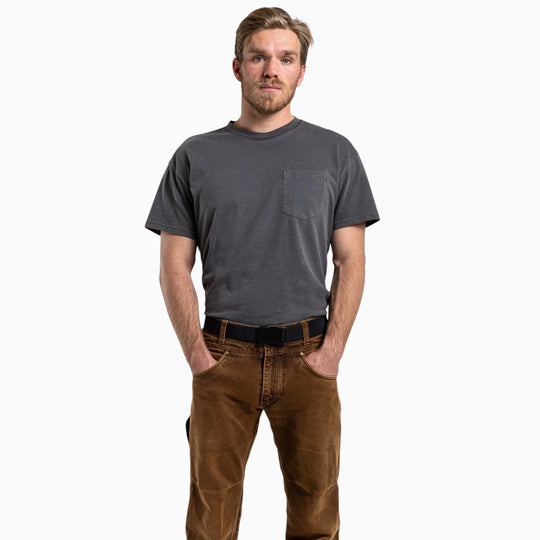 A man with short light hair, a beard, and mustache stands with hands in pockets against a plain white background, wearing a gray t-shirt and brown pants, radiating an air of dependable strength reminiscent of the Arcade Belts Utility Kit.