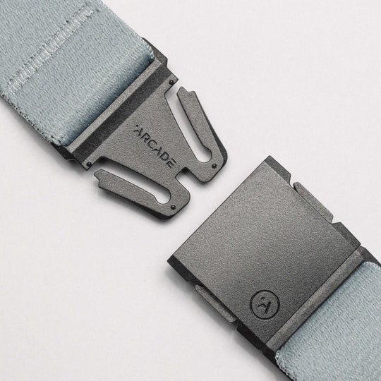 Close-up of a light blue stretch webbing belt from the Island Collection by Arcade Belts, featuring a black plastic buckle with ARCADE engraved on one side and a logo on the other, shown partially unbuckled on a white background.