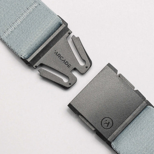 Close-up of a light blue stretch webbing belt from the Island Collection by Arcade Belts, featuring a black plastic buckle with ARCADE engraved on one side and a logo on the other, shown partially unbuckled on a white background.