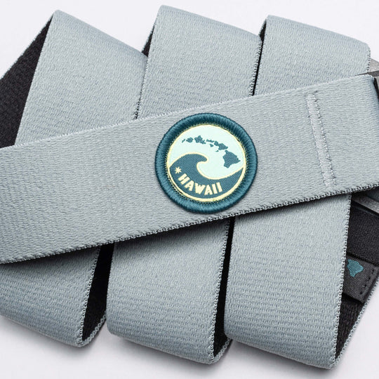 A light gray Arcade Belts Island Collection belt with elastic webbing features a round patch showing a wave, Hawaiian Islands, and #HAWAII in yellow-green. The band is neatly folded on a plain white background.