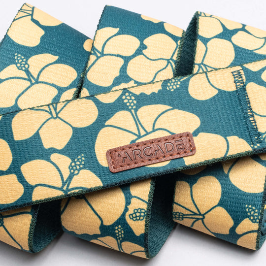 A teal elastic belt from Arcade Belts’ Island Collection features yellow hibiscus flowers and a brown leather patch labeled "ARCADE," coiled on a white background to capture the Aloha spirit of Hawaii.
