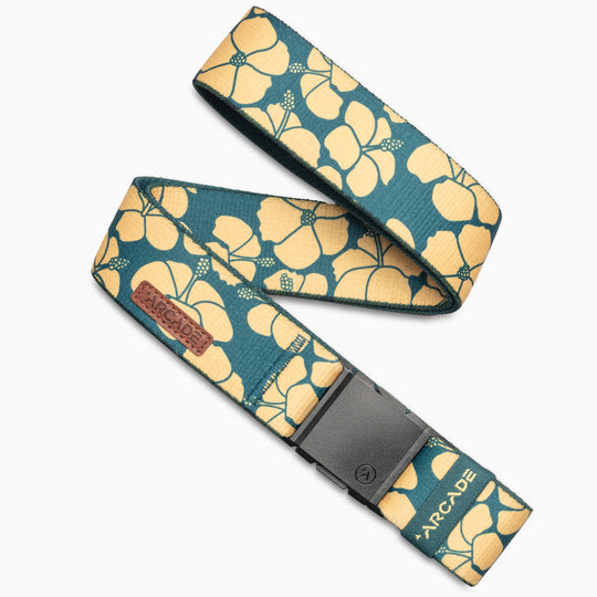 A teal fabric belt from Arcade Belts' Island Collection features a yellow hibiscus print, black rectangular buckle, and brown leather patch with "ARCADE," evoking the Aloha spirit of Hawaii in a loose looped display.