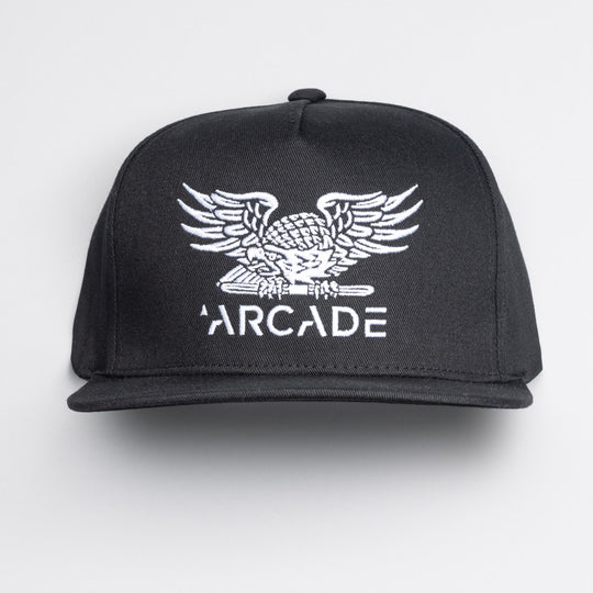 The Arcade High Crown Hat by Arcade Belts is a black snapback featuring a white embroidered eagle above ARCADE on the front, inspired by the brand’s iconic travel belts and stretch webbing. Photographed on a plain white background.