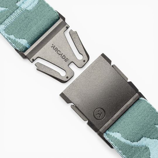 A close-up of a black metal belt buckle featuring the Arcade Belts logo, attached to the High Fives Duck Camo—a light blue and white patterned fabric belt—shown unclasped; a portion of proceeds from this product supports athletes via the High Fives Foundation.