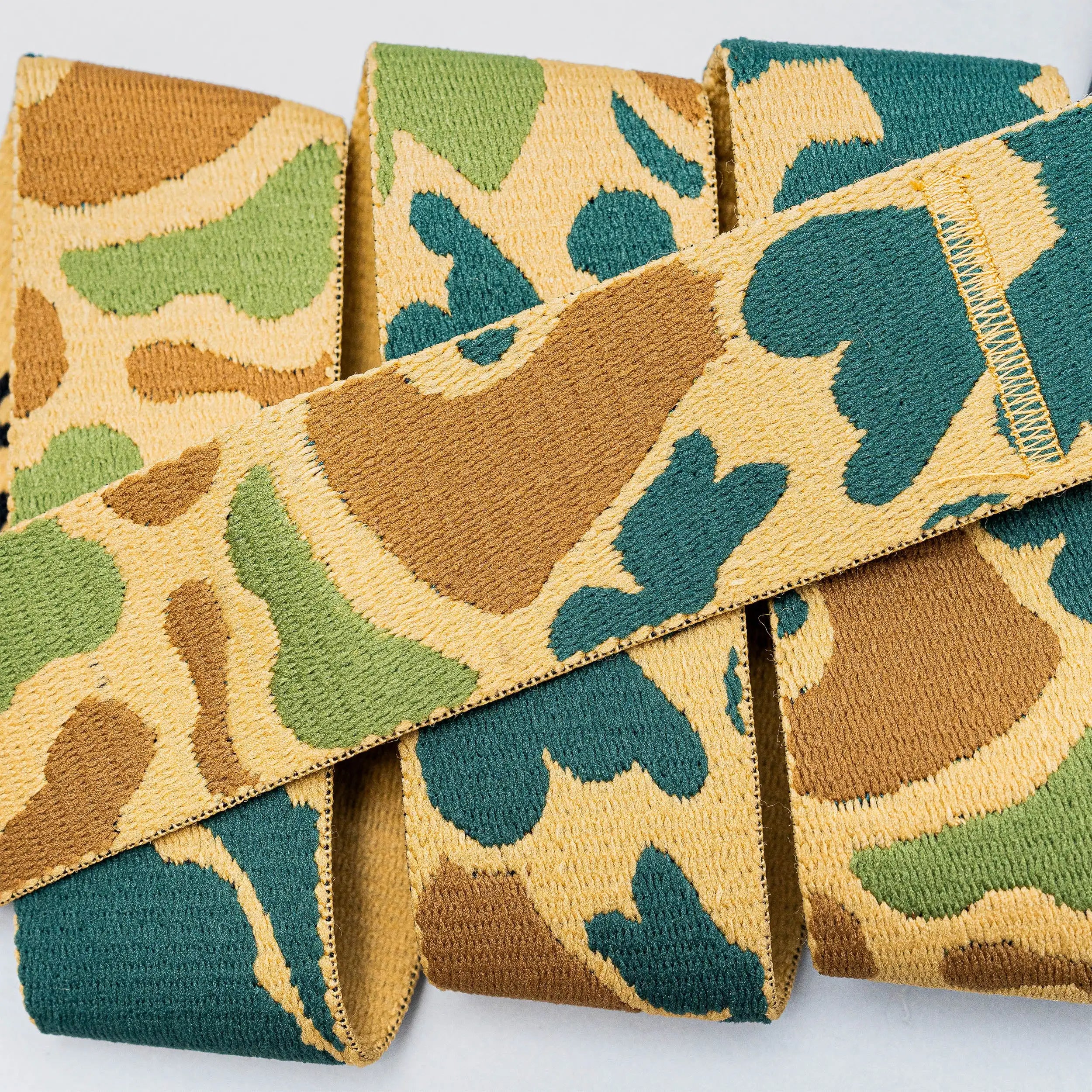 A close-up of High Fives Duck Camo fabric ribbon by Arcade Belts, featuring green, tan, and brown camouflage resembling stretch webbing, displayed on a light background.