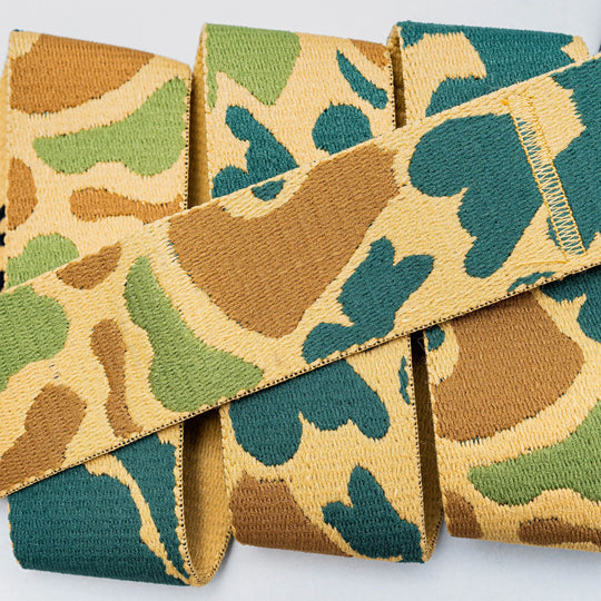A close-up of High Fives Duck Camo fabric ribbon by Arcade Belts, featuring green, tan, and brown camouflage resembling stretch webbing, displayed on a light background.