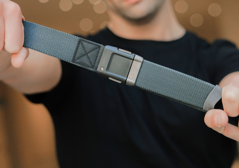 A person in a black shirt holds out an Arcade Belt with a black plastic buckle, stretching its gray stretch webbing toward the camera. The background is softly blurred with warm lights.